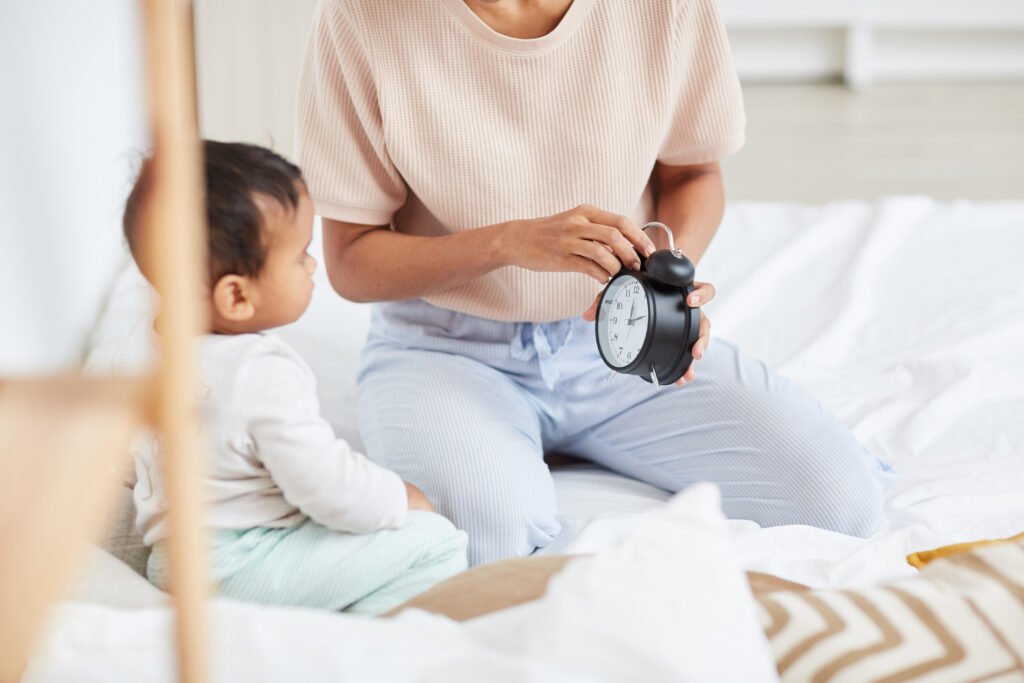 Close-up of mother holding alarm clock and checking the time with baby sitting on the bed and looking at clock